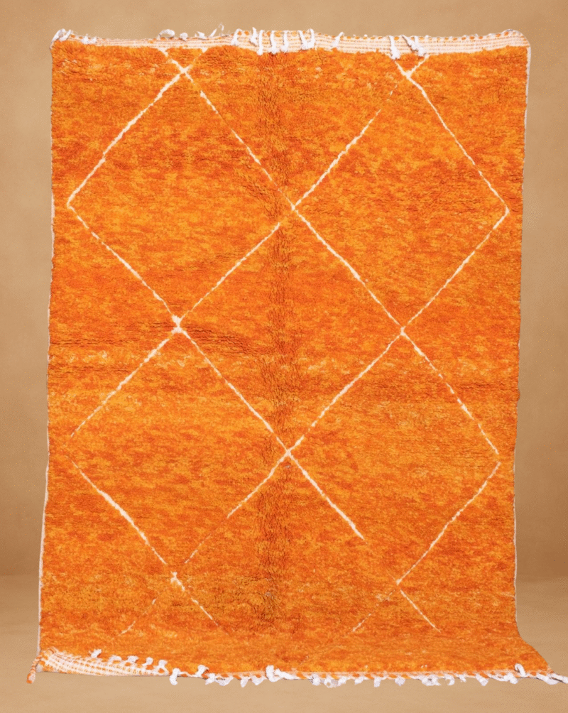 Handwoven Moroccan rug in vibrant orange with a subtle white diamond lattice pattern, displayed vertically against a neutral background. The rug features a soft, plush texture and fringes at the top and bottom edges.