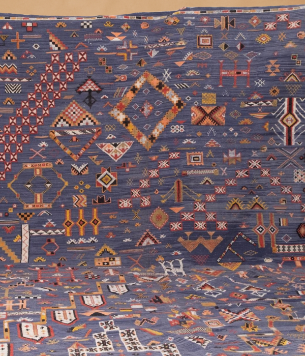 Kilim tribal rug with a rich indigo-blue base adorned with intricate, multicolored geometric and symbolic motifs in shades of red, yellow, orange, and white. The flat-weave textile showcases a dense arrangement of traditional Berber and tribal patterns, laid flat and cascading vertically against a warm neutral background.