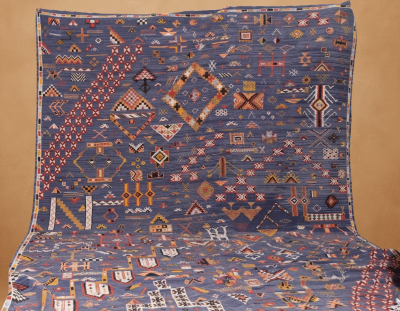 Kilim tribal rug with a rich indigo-blue base adorned with intricate, multicolored geometric and symbolic motifs in shades of red, yellow, orange, and white. The flat-weave textile showcases a dense arrangement of traditional Berber and tribal patterns, laid flat and cascading vertically against a warm neutral background.