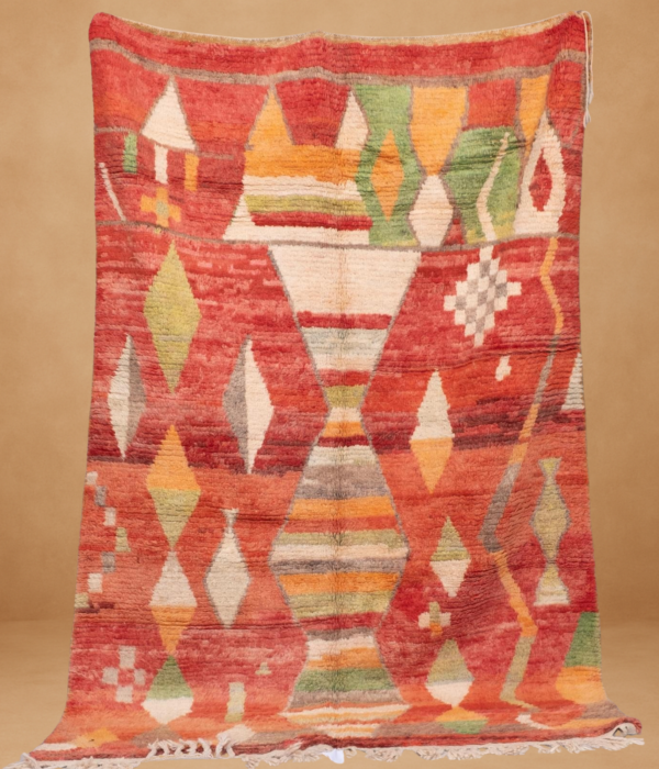 Moroccan Berber rug with vibrant geometric patterns on a red background, featuring diamonds, triangles, and abstract shapes in shades of green, yellow, orange, and white. Displayed vertically with white fringes at the bottom against a neutral beige backdrop.
