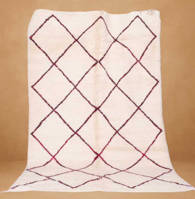 Beni Ourain rug featuring a soft ivory background with a traditional diamond lattice pattern in deep burgundy and subtle pink tones. Handwoven from natural wool, the rug is displayed vertically with a plush texture and minimal fringe, set against a warm neutral backdrop.
