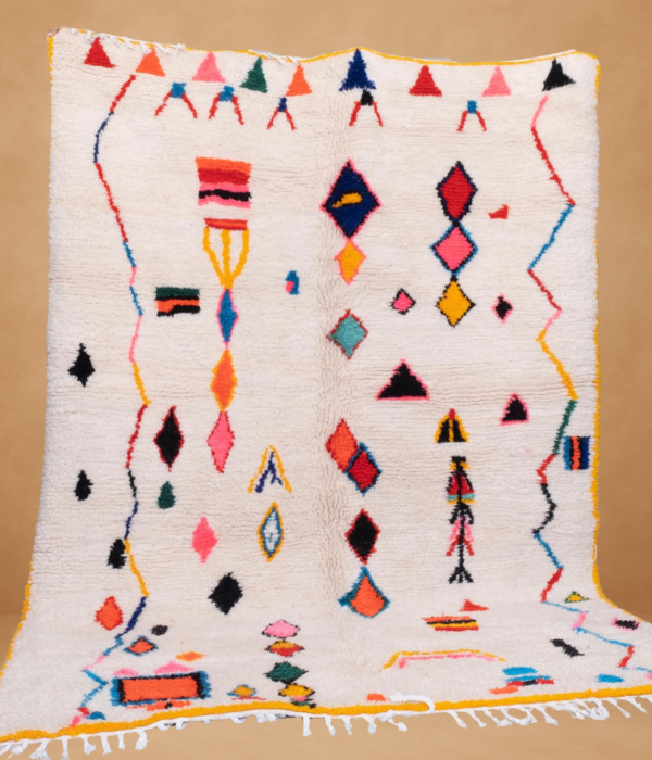 Moroccan Berber rug with a white base featuring colorful abstract and geometric motifs in shades of red, blue, pink, yellow, and black. The rug is displayed vertically with yellow edges and white fringes at the bottom, set against a neutral beige background
