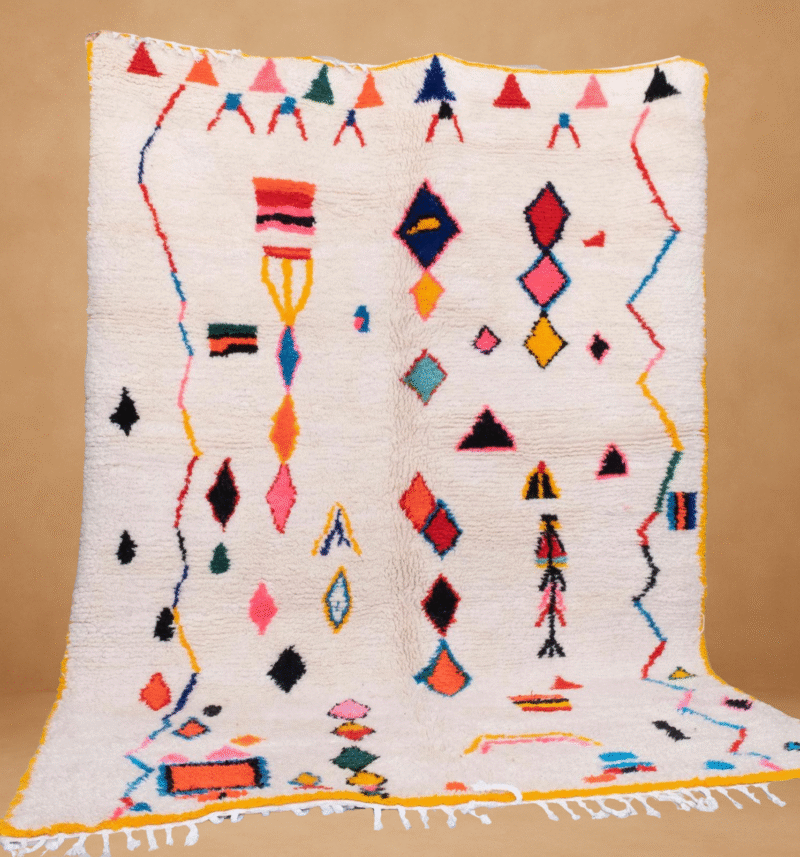 Moroccan Berber rug with a white base featuring colorful abstract and geometric motifs in shades of red, blue, pink, yellow, and black. The rug is displayed vertically with yellow edges and white fringes at the bottom, set against a neutral beige background