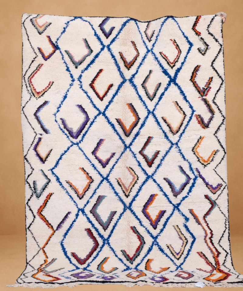 Beni Ouarain rug with a cream-colored wool base and a repeating diamond pattern outlined in blue, filled with multicolored V-shaped accents in shades of orange, purple, black, and red. The handwoven texture and vibrant geometry give it a lively yet traditional Berber feel. Displayed vertically with fringe along the bottom against a neutral background.