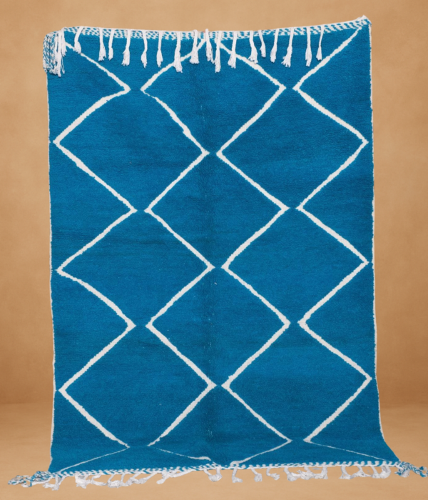 Beni Ouarain rug with a bold turquoise blue background featuring a classic white diamond lattice pattern. Handwoven from wool, the rug is displayed vertically with braided white fringes at the top and bottom, set against a warm neutral background.