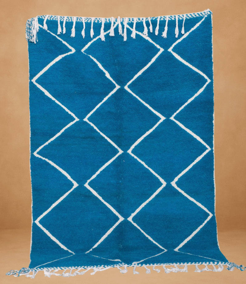 Beni Ouarain rug with a bold turquoise blue background featuring a classic white diamond lattice pattern. Handwoven from wool, the rug is displayed vertically with braided white fringes at the top and bottom, set against a warm neutral background.