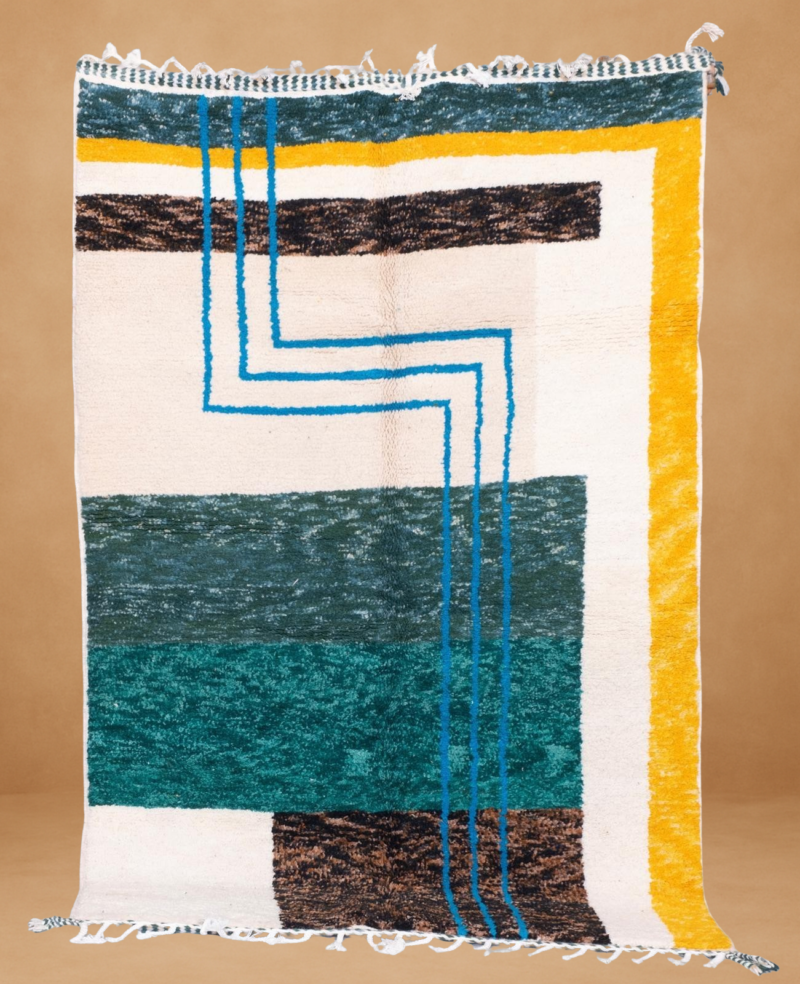 Modern Boujad rug with bold geometric blocks in forest green, teal, mustard yellow, and brown, accented by three vertical blue lines forming a right-angled pattern. Set on a cream background with textured wool, the rug is shown vertically with white fringes and a warm beige backdrop.