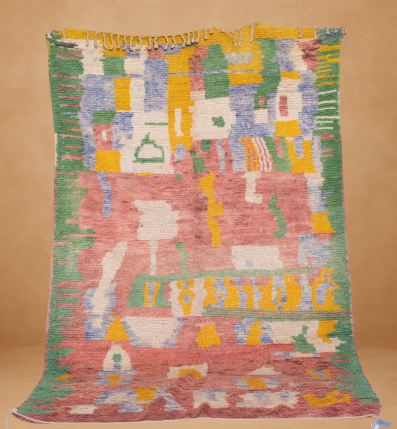 Playful Boujad rug with an abstract, painterly design in soft tones of rose, green, yellow, blue, and white. The composition features irregular blocks and freeform shapes, evoking a whimsical, mosaic-like pattern. Handwoven in wool and displayed vertically with short yellow fringe at the top, set against a warm neutral background.